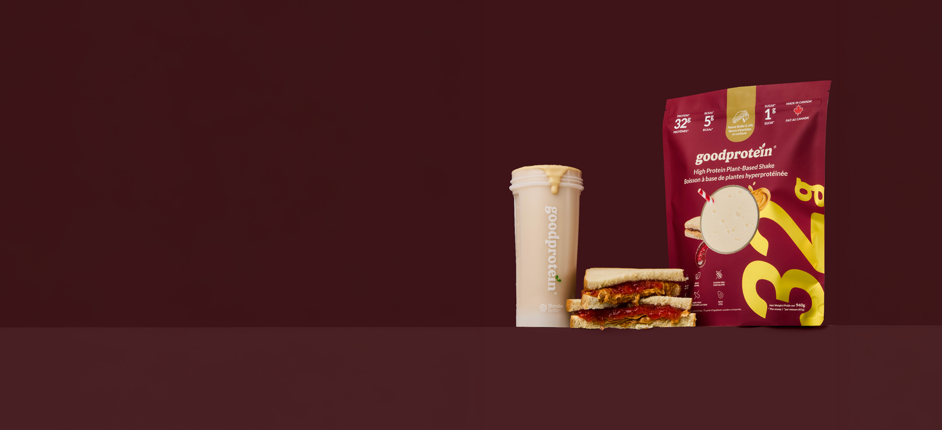 A Good Protein meal replacement pouch in dark red and yellow stands next to a protein shake cup and a sandwich on a dark burgundy background.