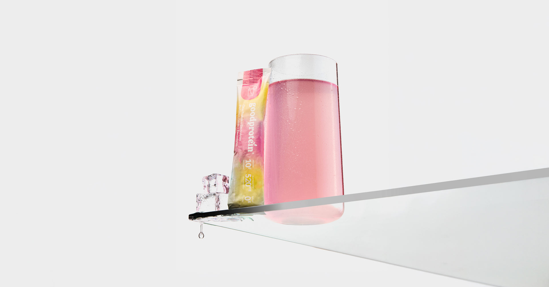 A glass of pink beverage with a colorful packet standing beside it on a reflective surface against a clean white background.