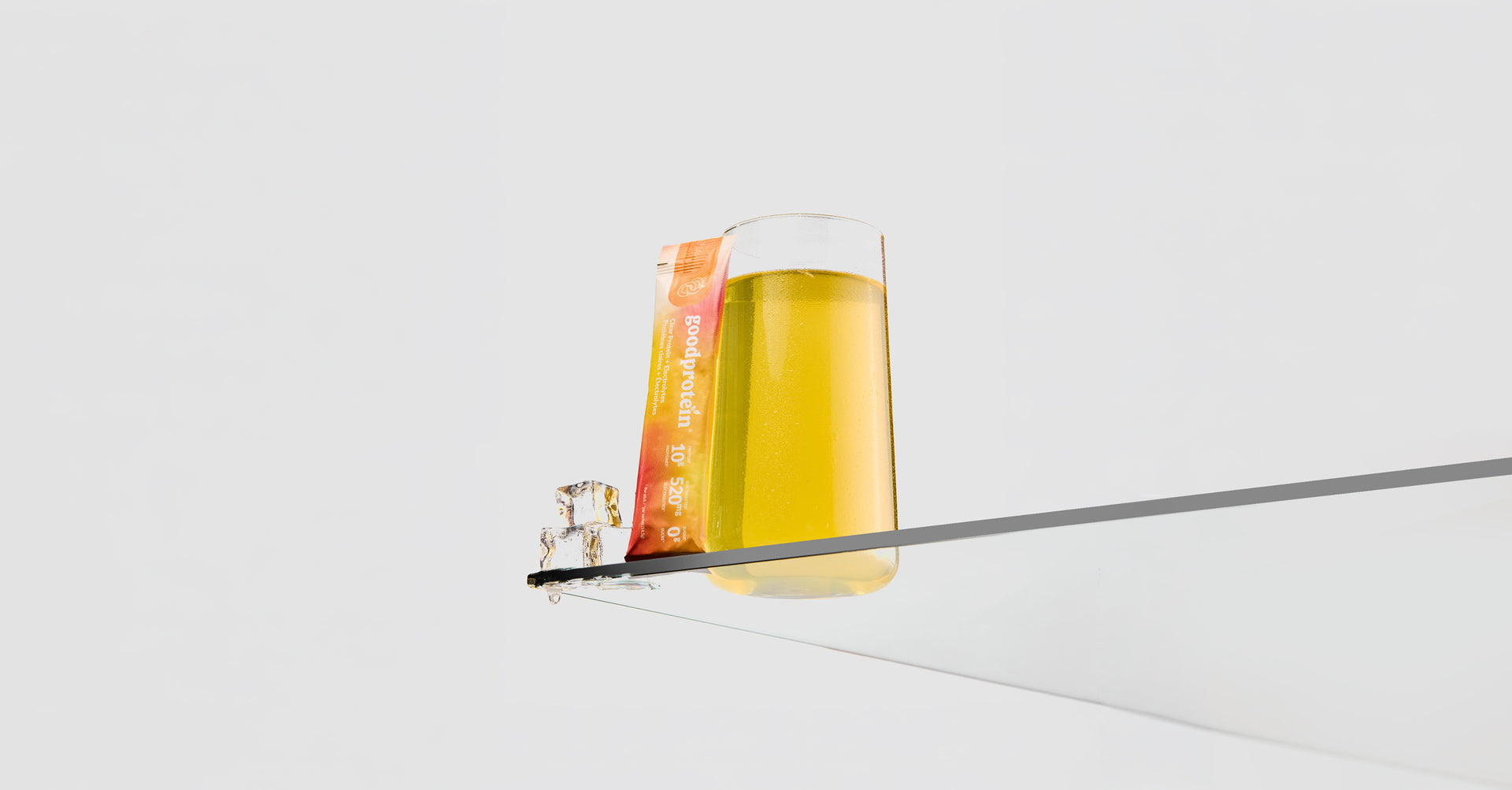 A clear cylindrical glass container filled with golden yellow liquid, shown resting on a thin gray bar against a clean white background. A small orange-and-white label is attached to the side of the glass.