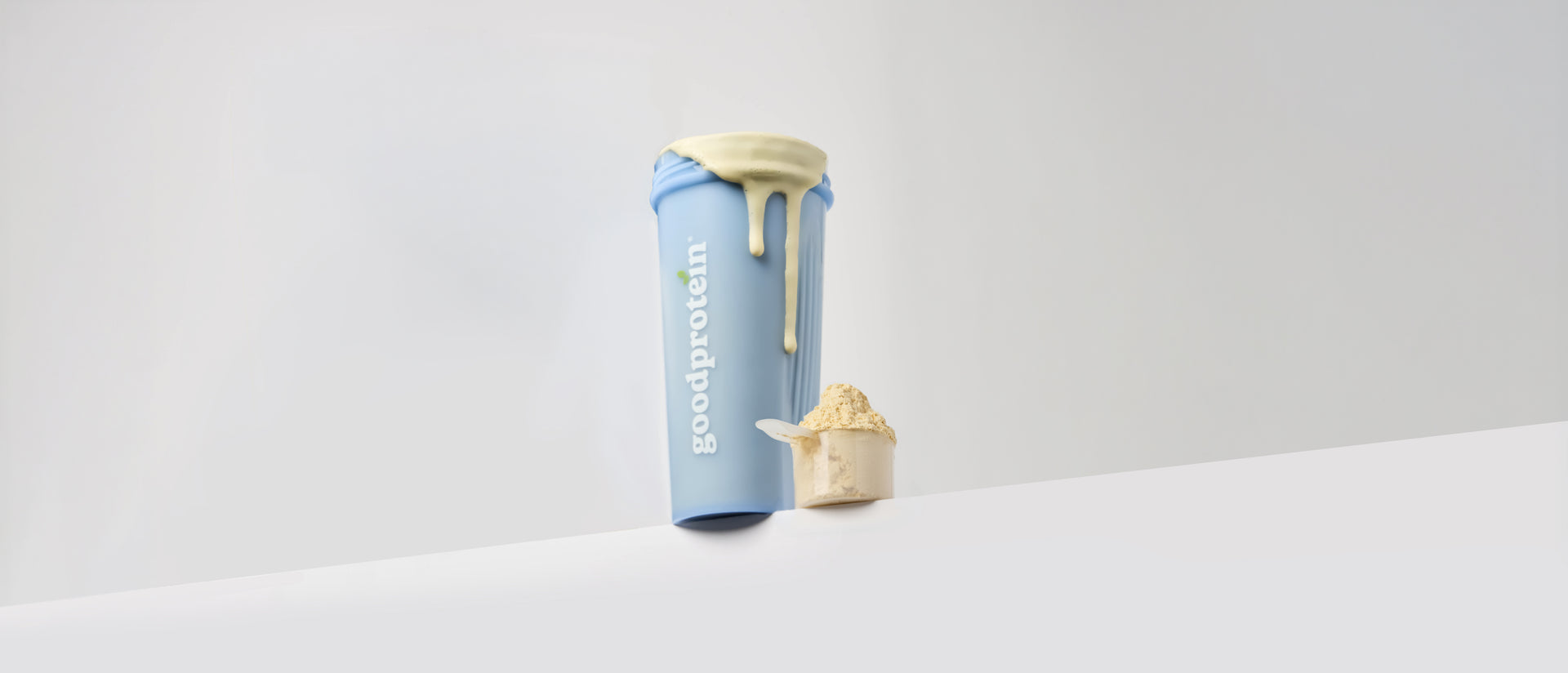 A light blue Goodprotein shaker bottle with a beige lid and a small beige scoop, set against a clean white and light gray background.