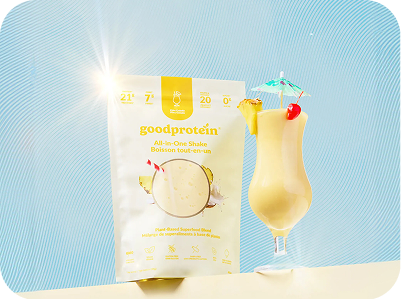 A bag of Good Protein All-In-One Shake powder stands next to a tall glass filled with a creamy yellow shake and garnished with a small umbrella and cherry, on a light blue background.