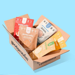 A cardboard box filled with multiple Good Protein powder pouches and stick packs in assorted flavors, photographed on a light blue background.