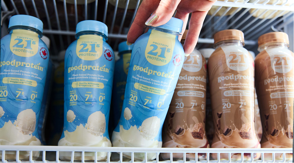 A hand reaching for a bottle of 21 Protein Good Protein powder on a refrigerator shelf, with blue and brown protein shake bottles visible.