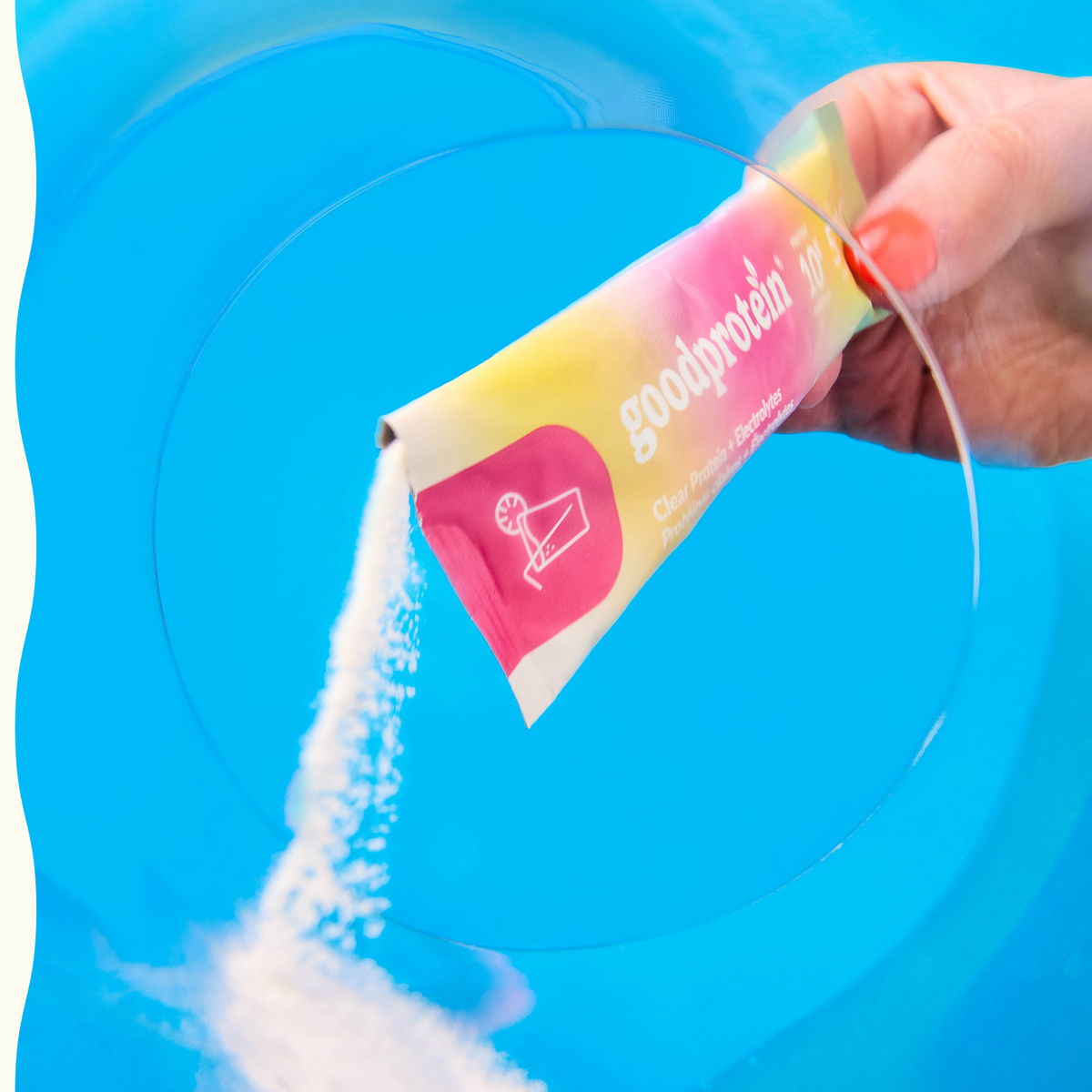 A hand pours white powder from a Goodprotein clay powder supplement sachet into a clear glass bowl, set against a bright blue background.
