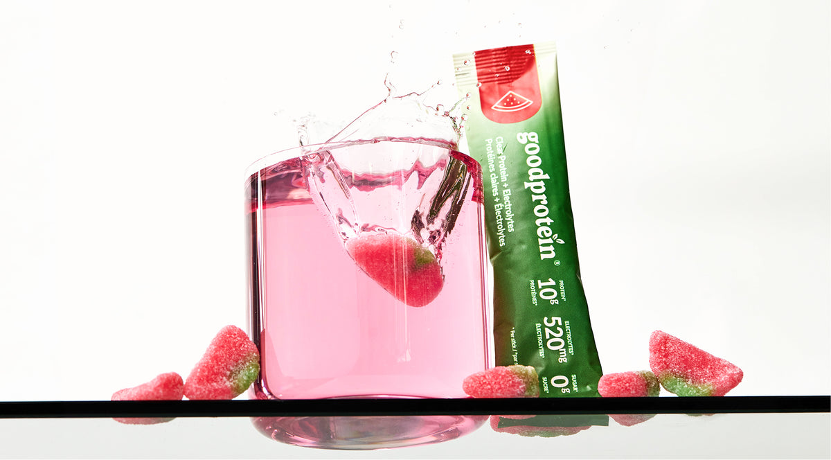 A single packet of GoodProtein protein powder stands next to a clear glass filled with pink liquid and a strawberry, with gummy strawberries scattered around on a reflective black surface against a white background.