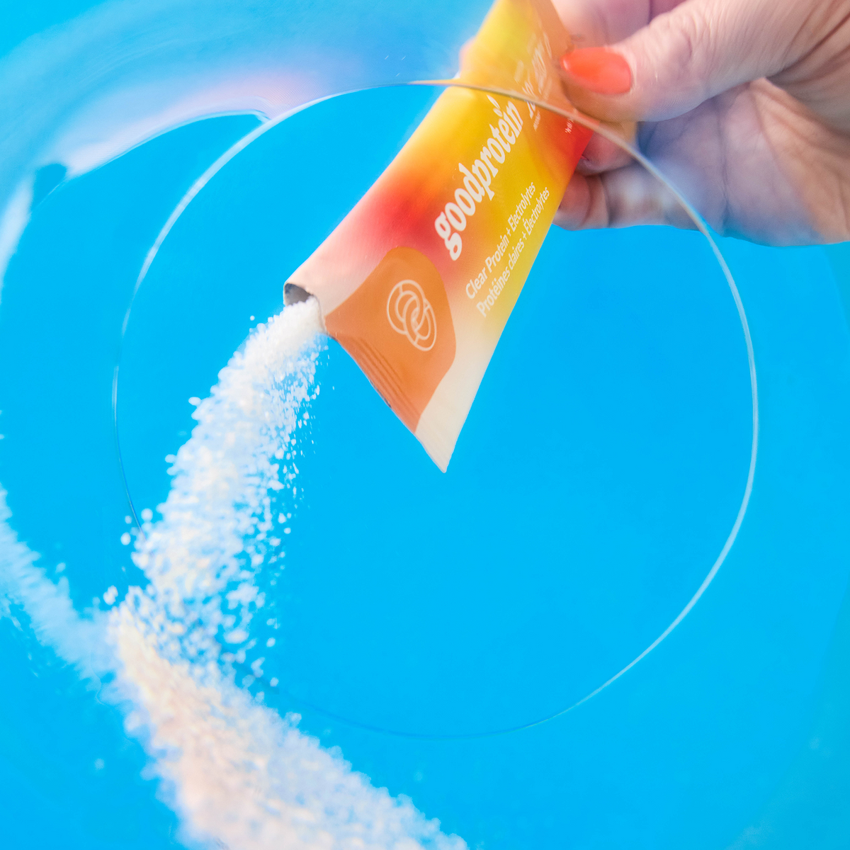A hand pours white powder from an orange and yellow Goodprotein sachet into a clear circular dish over a bright blue background.