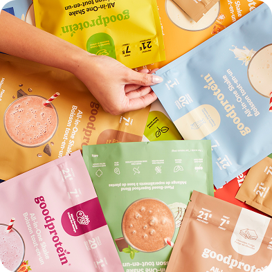A hand is shown holding a light blue Goodprotein All-in-One Shake pouch, surrounded by other colorful pouches in yellow, green, pink, and brown on a patterned background.